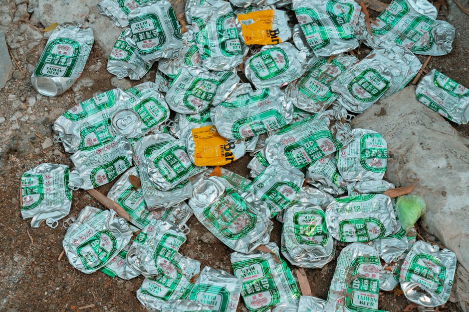 A close-up view of numerous crushed aluminium cans scattered on a dirt ground, with some cans displaying green and white labels and others featuring orange and yellow labels. The cans, which are weathered and crumpled, appear to be discarded waste likely originating from beverages such as beer or soft drinks. They are scattered randomly among small pieces of debris, dried leaves, and natural dust, indicating an outdoor environment possibly on a pavement or garden area. The metallic surfaces of the cans reflect ambient light, highlighting their crumpled textures and varying levels of cleanliness. Some cans are partially hidden beneath others, creating a layered effect, while a few are positioned next to a large white plastic bag and nearby dirt patches. This scene depicts typical household or outdoor waste, relevant to private rubbish collection and alternative waste handling services, such as those offered by Rubbish Clearance Barnes, which specialize in managing bulky or small-scale debris like these cans in an efficient and environmentally responsible manner.