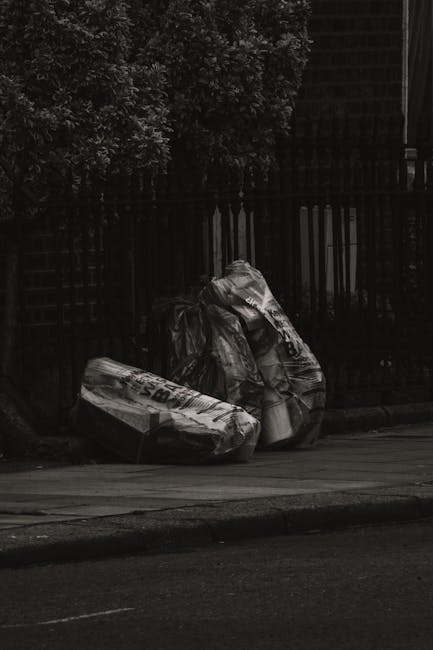 Two large garbage bags, made of black plastic with visible creases and wrinkles, are placed on the pavement near a black metal fence with vertical bars. The bags appear to contain household waste or discarded materials and are positioned close to each other, leaning slightly to the right. The background features an arrangement of dense, leafy greenery and a dark, possibly brick or wooden, structure behind the fence. The scene is outdoors, on a street or sidewalk, with natural lighting casting subtle shadows. The overall setting suggests a private or communal rubbish collection point, where residents or service providers like Rubbish Clearance Barnes might conduct either independent waste handling or preparatory rubbish removal for collection, consistent with their services focused on quick and efficient clearance on sites such as Barnes High Street.