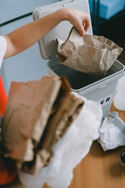 A person is seen disposing of paper waste into a small, grey recycling bin or rubbish container, with their right hand holding an empty paper bag above the open lid. The person's arm is visible, and they appear to be placing the bag into the container, which is situated on a wooden surface. In the foreground, there are additional crumpled paper bags, some slightly blurred, indicating active waste separation. The background includes a portion of an indoor environment, with part of a window or frame visible, suggesting this scene might relate to private waste handling or at-home rubbish disposal. The overall setting emphasizes organized waste sorting, aligning with private rubbish collection practices such as those managed by Rubbish Clearance Barnes, supporting environmentally responsible waste removal options outside of local authority collection methods.