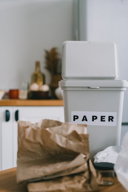 A person is seen disposing of paper waste into a small, grey recycling bin or rubbish container, with their right hand holding an empty paper bag above the open lid. The person's arm is visible, and they appear to be placing the bag into the container, which is situated on a wooden surface. In the foreground, there are additional crumpled paper bags, some slightly blurred, indicating active waste separation. The background includes a portion of an indoor environment, with part of a window or frame visible, suggesting this scene might relate to private waste handling or at-home rubbish disposal. The overall setting emphasizes organized waste sorting, aligning with private rubbish collection practices such as those managed by Rubbish Clearance Barnes, supporting environmentally responsible waste removal options outside of local authority collection methods.