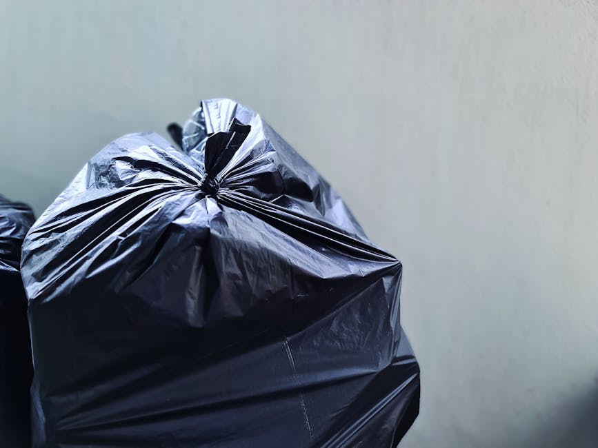 A close-up view of several black plastic rubbish bags filled with waste, tightly knotted at the top, stacked against a plain, light-colored wall. The bags have a glossy surface with visible creases and folds, indicating they are full and ready for disposal. The setting appears to be indoor or in an outside storage area, with natural or diffused lighting illuminating the scene. These bags, common in private waste handling and rubbish clearance services, are positioned in the foreground, while the background remains minimal and unobstructed. This image exemplifies typical waste collection practices where rubbish clearance companies like Rubbish Clearance Barnes facilitate alternative waste disposal options, such as on-site collection or private rubbish removal, rather than relying solely on local authority bins or council services.