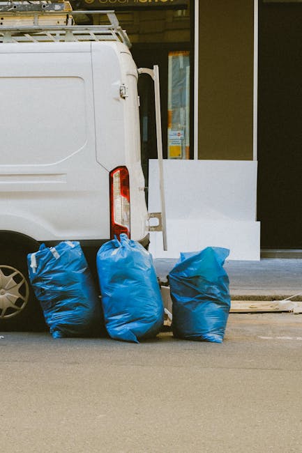 The image shows the rear side of a white cargo van parked on a paved street in front of a building with a large window and brown awning. Four blue rubbish bags, made of thick plastic with a slightly shiny surface, are placed on the ground, leaning against the back of the van. The bags appear sealed and full, with some creases and folds visible on their surfaces. Behind the bags, part of a metal bumper and the van's taillight are visible. The building in the background features a glass window reflecting the street scene, with a brown awning above the entrance. The scene is evenly lit, suggesting natural daylight, and the overall setting indicates an instance of private waste disposal, likely arranged through a private rubbish removal service such as Rubbish Clearance Barnes, supporting the broader topic of independent rubbish collection and on-site clearance options in the UK.