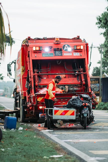 A street scene showing a waste collection operation with a red rubbish truck parked on the roadside. A worker wearing a high-visibility yellow and red uniform is positioned behind the truck, operating the mechanical arm or hatch at the rear of the vehicle. The back of the truck is open, revealing its internal components, and there is a large black plastic bag placed on the back platform. The scene includes a motorbike parked nearby, partially obscured by the worker. In the background, there are trees, power lines, and a signboard for a local business, suggesting a commercial or mixed-use area. The surroundings display a paved road with a designated parking or curb zone, and some litter or debris on the ground near the worker and the vehicle, indicating ongoing waste collection or rubbish removal activities. The lighting appears natural, typical of an overcast day, emphasizing the professional and neutral tone of waste management operations, consistent with the services provided by Rubbish Clearance Barnes for private rubbish removal and on-site clearance in the area.